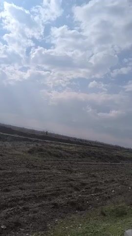 The video shows a landscape scene under a cloudy sky. A field of dark soil occupies the lower portion of the frame, and beyond it, there is a slightly elevated area covered with grass and sparse vegetation. A road or highway is visible in the distance, with vehicles moving along it. The sun is partially obscured by the clouds, creating a diffused, bright light.