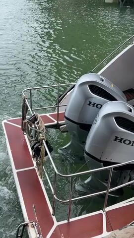 The video shows the rear of a boat as it travels through the water. Two silver Honda outboard motors are visible mounted on the stern. The water behind the boat is churning with white foam and waves, indicating motion. The boat has a maroon deck with white trim, and stainless steel railings. Ropes are tied to a railing on the deck. The water is a greenish-gray color, and the sky appears overcast.