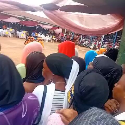A video shows a crowd of people gathered outdoors, mostly women, under a large tent with red and pink fabric draped overhead. Many of the women wear head coverings. They are standing close together, looking in the same direction, suggesting they are watching an event or listening to a speaker. In the background, there are more people seated at tables under additional tents. A building with a brown roof is visible in the distance. The ground is dirt.
