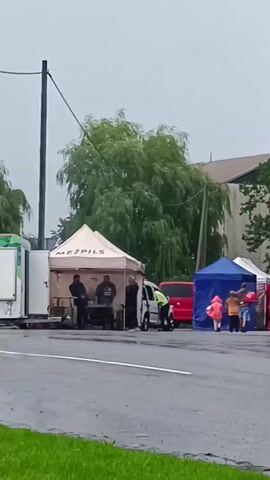 The video shows an outdoor event under overcast skies. In the foreground, the rear of a red car is visible. Behind it, a wet asphalt road leads to a row of tents and stalls. To the left, a white tent labeled "MEŽPILS" has several people standing around it, with what appears to be a large black pot or container. A red van is parked behind this tent. Further to the right, a blue tent has a few children in colorful jackets and adults gathered. Next to the blue tent is a red tent with a clear plastic enclosure. Two large feather flags are prominently displayed: one white with text, and a red one with a white symbol, possibly a cross. More people are seen standing near these flags and tents, some interacting with a white portable unit. A black SUV is parked behind the stalls on the right. The overall environment suggests a community gathering or market, possibly affected by rain given the wet road and overcast conditions.