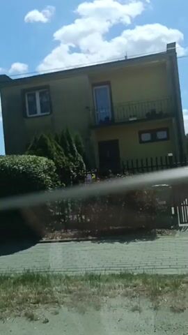 The video shows a view from a moving vehicle, likely a car, looking out through the windshield. The exterior environment is characterized by a bright, sunny day with scattered clouds. On the right side of the frame, a two-story building with a light-colored facade and white-framed windows is visible. Trees, including a large evergreen, and a fence are also present. The foreground shows a paved surface, possibly a driveway or sidewalk, with some grass and weeds growing between the paving stones. The camera is in motion, causing a slight blur and a horizontal line across the frame, which appears to be part of the vehicle's structure, possibly a window frame or wiper. The footage is brief, lasting only a few seconds, and captures a fleeting glimpse of the surroundings.