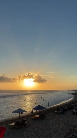 This is a vertical, eye-level video of a beach at sunset. The sun is partially obscured by clouds and is setting over the ocean. There are waves in the water, and several people are swimming or surfing. On the beach, there are umbrellas, lounge chairs, and beanbag chairs. Some people are sitting in the chairs, and others are walking along the shore. The sky is a clear blue, with some light clouds. The overall mood is peaceful and relaxing.