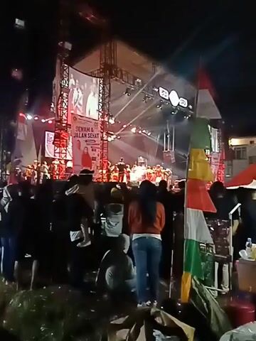 The video shows a nighttime outdoor event with a stage set up in the background.  A large crowd of people is gathered in front of the stage, many facing the performance. Lights are illuminating the stage, and a large screen is visible above it.  In the foreground, several individuals are visible, including a person in a red jacket and jeans, and another person in a green shirt.  A table with various items, possibly drinks and snacks, is set up to the right, covered by a red umbrella.  Indonesian flags are displayed on a pole to the left of the table.  The overall atmosphere appears to be a concert or public gathering.