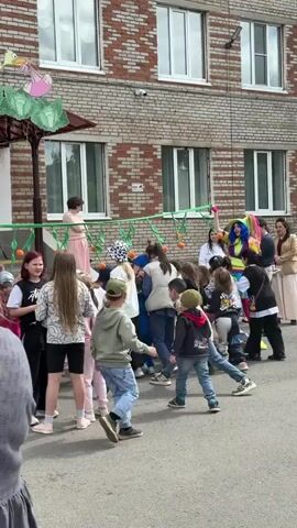 Outside a two-story brick building, children are participating in a limbo game. An adult in a colorful clown costume holds one end of a green limbo stick with orange balls attached, while an unseen person holds the other end. The children line up and duck under the stick, cheered on by adults and music playing from a speaker set up in front of the building’s entrance. People in the foreground are watching and filming the event with their cell phones. Decorations are on the building’s roof. The building appears to be a school or community center, based on the signage and exterior decor.