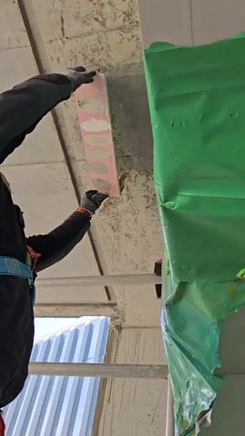 A worker on a ladder, wearing a safety harness and gloves, holds up a pink board with different shades of grey and white materials attached to it, against a rough concrete ceiling. Some text is written under each material swatch. A green plastic sheet hangs nearby. The setting appears to be a construction site with visible building structures and an outside view of a blue container through an opening. The worker seems to be comparing the swatches to the ceiling's color.
