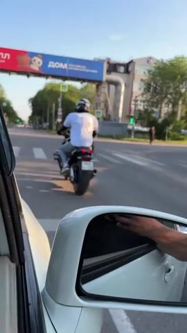 The video shows a view from inside a car, looking out at a street scene. In the foreground, the side mirror of the car is visible, reflecting a person's arm and hand. In the middle ground, a person on a motorcycle is riding away from the camera, crossing a pedestrian crossing. Behind the motorcycle, there is a large billboard overhead with text and images, and a building on the right. The environment appears to be an urban street during the day with traffic lights and pedestrian crossing signs visible. The sky is blue with some clouds, suggesting pleasant weather.