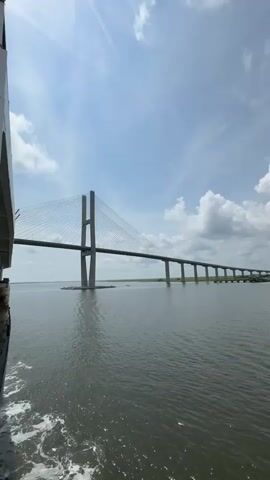 A long shot captures a large suspension bridge spanning a body of water under a cloudy sky. The camera is positioned on a vessel, with the edge of the ship visible in the lower left corner, showing churning water from its movement. The bridge itself features two tall concrete towers connected by a roadway, supported by numerous cables. The water is calm, with a few small boats in the distance. The shoreline is visible on the horizon, lined with green vegetation. The lighting suggests it is daytime, likely midday.