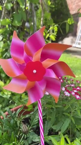 A colorful pinwheel spins in a flower pot in a backyard. The pinwheel is made of pink and orange petals and has a red center with a white button. The pinwheel is attached to a pink and white striped stick. The flower pot is filled with green plants and pink flowers. The backyard has a green lawn, a brick house, and some trees. A white step stool is visible in the background.