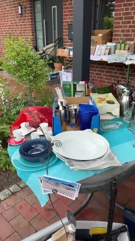 The video captures an outdoor market scene, likely a garage sale or flea market, set up under a covered patio with a brick wall and red brick-paved ground. Two tables and a clothing rack display various items. The left table, covered by a light blue cloth, holds white plates, blue bowls, glassware, and cutlery containers, with a sign reading "Alles 1€" (Everything 1€) behind it. The right table, a smaller foldable metal type, features books, children's toys, and framed items. A silver metal clothing rack to the right displays assorted garments, with folded textiles stacked beneath. Several people browse the items: a person in a dark jacket arranges goods on both tables, a woman in a dark hoodie and blue jeans stands with a small child near the right table, and a woman with blonde hair walks past with a young girl. Another individual with a red backpack approaches the clothing rack. Blue rubber boots and a box of books are visible on the ground.