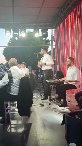 A group of musicians is performing on an outdoor stage. The stage is lit with bright spotlights and has a backdrop of red, vertical streamers. In the foreground, a man in a white shirt and black pants is playing an accordion. To his right, another man in a white shirt and black pants is playing a keyboard with drumsticks. To the right of the keyboard player, a man in a white shirt and black pants is playing a guitar. A lead singer, also in a white shirt and black pants, stands center stage, holding a microphone and a small percussion instrument. Several other people are visible in the background, some seated and some standing. The overall atmosphere appears to be festive.