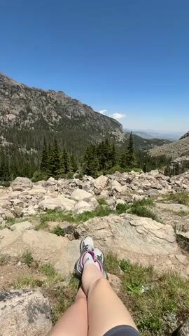 The video shows a person's legs and feet, clad in hiking shoes, resting on a rocky, grassy slope. The person is looking out at a mountainous landscape under a clear blue sky. The terrain is rugged, with large rocks and scattered pine trees. In the distance, a valley opens up, revealing more mountains and a hint of water, possibly a lake. The camera pans slightly, offering different perspectives of the scenic view.
