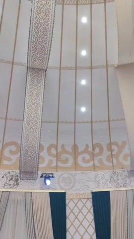 The video shows an upward view of the interior of a large, circular structure, possibly a yurt or a domed tent. The walls are covered with fabric panels featuring a pattern of vertical lines and decorative borders. There are multiple small, circular lights embedded in the ceiling. Two decorative fabric hangings, one with a geometric pattern and another plain beige fabric, descend from the ceiling. The camera slowly pans upwards, revealing the dome shape and the intricate patterns on the central part of the ceiling, which appears to be a decorative fabric disc with various motifs. At the very bottom of the frame, there are glimpses of what appears to be decorative drapes or curtains, and a spotlight is visible. The overall impression is of an ornate and culturally inspired interior space.