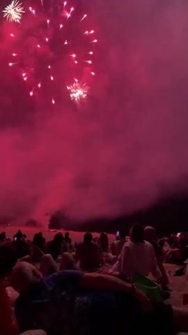 A fireworks display unfolds over a beach, with a crowd of people seated on the sand watching. The sky is filled with bursts of red, green, and gold fireworks, creating a smoky haze. The people are silhouetted against the bright explosions, and some are holding up their phones to capture the event. The overall atmosphere is one of celebration and awe.
