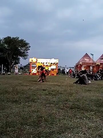 The video shows an outdoor performance, possibly a traditional dance or martial arts demonstration, on a grassy field under an overcast sky. In the foreground, two individuals are engaged in a physical interaction, with one appearing to be on the ground and the other standing over them, reaching down. They are both wearing traditional attire that includes patterned fabrics and dark tops. Behind them, a brightly lit, orange and yellow stage or booth with the number "76" prominently displayed is visible. Several other people, likely spectators, are seated or standing in the background, some near tents and structures. The overall atmosphere suggests a cultural event or festival.