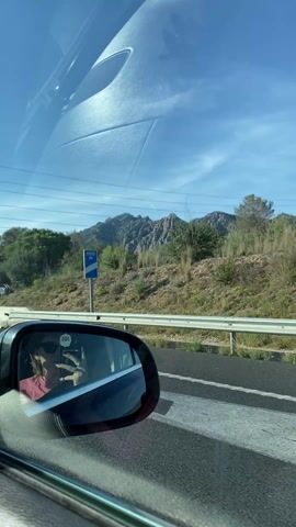 The video shows a car traveling on a highway. The view is from the driver's seat, showing the road ahead, a truck in front of the car, and road signs indicating directions to Barcelona and Esparreguera. A small, gold-colored statue of a dog is on the dashboard. The side mirror reflects the passenger, who is holding a phone and possibly taking a photo or video. Outside the car, there are trees and a mountain range visible in the distance under a blue sky with some clouds. The car is moving along a concrete barrier.