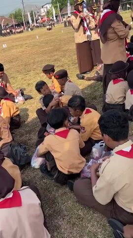 A group of children dressed in scout uniforms, some wearing hijabs, sit on a grassy field. They appear to be having a break, with some eating or looking at food items. In the background, other scouts are lined up and a few adults are visible. The environment appears to be an outdoor event or gathering.