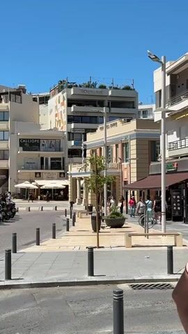 A street scene on a sunny day with a clear blue sky. The foreground shows a paved area with bollards and a street lamp. A small tan car is parked in the middle ground. Beyond the car, there are trees with lush green foliage. Further back, buildings with shops and cafes line the street. A row of parked motorcycles is visible on the left. People are walking and sitting in the background. One man in a black t-shirt is walking towards the camera in the foreground.