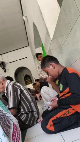 The video shows several people, predominantly young males and one older male, sitting on the floor of what appears to be a mosque. The lighting is dim, suggesting an indoor setting. The people are dressed in casual clothing, some wearing Islamic attire like prayer caps. One young boy in the foreground is looking down at something in his hands, possibly a phone or a prayer bead. Another younger boy stands behind him, looking towards the camera. An older man is seen to the left, also looking down. The environment features tiled walls and a high ceiling with visible lighting fixtures. The atmosphere appears to be one of prayer or quiet contemplation.