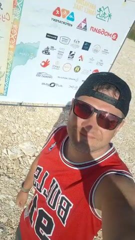 A man in a red "BULLS 23" basketball jersey, a black baseball cap worn backward, and red-tinted sunglasses is recording a selfie video. He has a slight smile and holds the camera, panning slowly to reveal the background. The scene is an outdoor event on a gravel-covered ground, under a clear sky with mountains visible in the distance. Various tents, including orange, red, blue, and green, are set up in the background. Numerous people are scattered throughout the area, walking and standing. Several cars are parked, and at least two dogs are seen roaming. In the initial frames, a large white banner with drawings of palm trees and mountains is visible behind the man. The man briefly raises his left fist at one point during the recording.