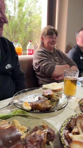 A group of people are sitting around a table laden with food and drinks. There are various dishes including meats, pastries, a ring-shaped cake with chocolate and almonds, and what appears to be a bottle of alcohol. A woman with a dog on her lap is seated in the background on a couch. Two other individuals are closer to the camera, one woman with her arms crossed and a man seated to her right. The room seems to be indoors, possibly a dining room, with natural light coming through a window. The overall atmosphere suggests a casual gathering or meal.