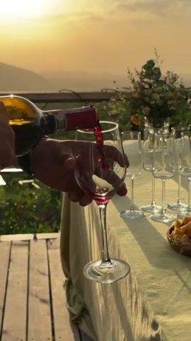 Against a golden sunset backdrop, a person is pouring red wine from a bottle into a stemmed glass. Several other wine glasses are lined up on a table covered with a pale green cloth, along with a basket containing cheese or bread. The event takes place outdoors, on a wooden deck overlooking a hazy landscape. The person is wearing a watch on their left wrist. A floral arrangement adds to the celebratory ambiance.