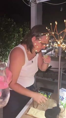 A woman with dark hair and glasses, wearing a white tank top and dark pants, is seen in a dimly lit outdoor setting at night. She is holding a lime wedge to her mouth and appears to be smelling it or possibly tasting it. To her left, a person's hand is visible holding a clear glass. In the background, there's a decorative light fixture resembling a starburst with many small lights, a table with some items on it including glasses and what appears to be a drink carton, and some foliage. The woman's facial expression suggests she might be experiencing a strong sensation, possibly from the lime. The overall scene suggests a social gathering or a casual moment outdoors in the evening.