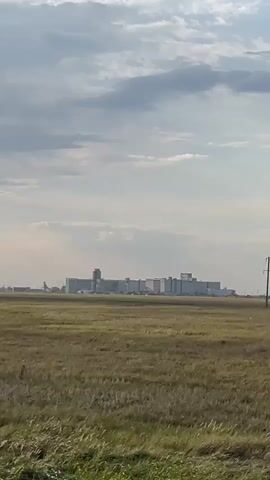 The video shows a wide, flat, grassy field under a cloudy sky. In the distance, a large, multi-building industrial complex is visible. The complex appears to be a series of connected structures with varying heights. To the left of the complex, an airplane is visible on the ground. Further to the right in the distance, numerous electrical transmission towers are scattered across the landscape. The grass in the foreground is dry and tan, suggesting a rural or agricultural setting. The camera is stationary, and the video appears to be shot from a distance.