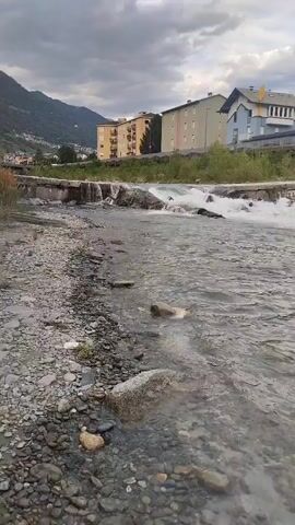 The video shows a body of water, likely a river or stream, flowing over rocks. On the left side of the frame is a rocky shore with various sizes of pebbles and stones. The water itself is clear and appears to be moving at a moderate pace. In the background, there are several buildings, appearing to be apartment complexes or residential buildings, situated on a gentle slope leading up to a mountain range. The sky is overcast with clouds, suggesting a subdued or potentially rainy day. The overall scene depicts a natural waterway in an urban or semi-urban setting.