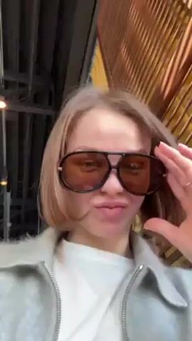 A woman with fair skin and short reddish blonde hair wearing a white top and gray zip up jacket removes large aviator style sunglasses with orange tinted lenses and places them to her right. She pauses, then puts on a pair of frameless sunglasses with gold arms and light blue lenses. The woman is inside a room with a slatted wood wall behind her and overhead exposed pipes and lights. A female with dark hair in a red hair tie is visible in the background to the left.