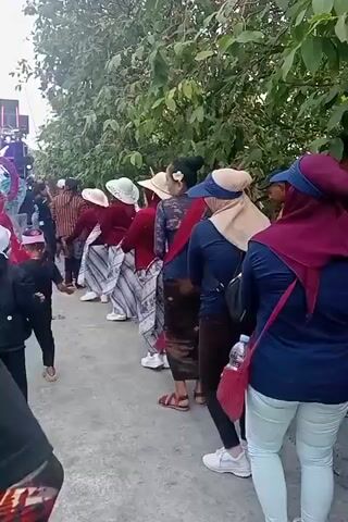 The video shows a group of people, primarily women, in a line dancing outdoors. They are all facing the same direction, with their arms extended forward. The people are dressed in casual clothing, with some wearing headscarves and hats. There are trees and bushes in the background, as well as some speakers and equipment on the left side of the frame. The ground is paved, and there is a fence on the right side. The lighting suggests it is daytime.