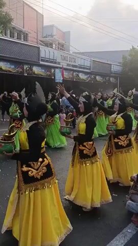 A group of women in yellow and green traditional costumes are dancing in a street. They are wearing headscarves and ornate headdresses. Some are holding bowls with decorative items inside. They appear to be part of a parade or cultural performance. In the background, there are buildings with signs, including one that says "TOKO DUTA". Spectators, including children, are visible on the side of the street. The lighting suggests it is daytime, possibly late afternoon.
