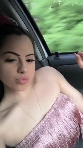 This video shows a young woman with dark hair and makeup, wearing a strapless pink top with fringe, posing in the passenger seat of a car. She is looking at the camera and puckering her lips. The background outside the car window shows a blur of green trees and foliage, indicating the car is in motion. The lighting suggests it is daytime. The woman appears to be relaxed and playful. There is no visible text in the video. The context provided indicates the location is Kichevo, Bulgaria.