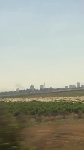 This is a landscape video taken from a moving vehicle, likely a train or car, in the Yoav Regional Council of Israel. The scene features agricultural fields in the foreground and mid-ground, with a city skyline in the distance.

In the mid-ground, there are rows of lush green crops, possibly vineyards or orchards, stretching across the frame. Some areas of the fields appear to be covered with netting, which could be for protection against pests or birds. The foreground consists of dry, brownish earth and some sparse vegetation, suggesting a more arid or recently harvested area.

The background is dominated by a hazy, pale blue sky, indicating a bright and likely warm day. A distant city skyline is visible on the horizon, characterized by several tall buildings and structures, including what appear to be electrical pylons. The buildings are silhouetted against the sky, making their exact details unclear.

The movement of the vehicle causes some motion blur in the foreground and mid-ground, creating streaks of color that emphasize the speed of the journey. The overall impression is one of a rural or semi-rural landscape transitioning towards an urban environment, captured under clear, bright daylight. There is no visible text in the video. The mood is neutral, observational, and perhaps evokes a sense of travel and distance.