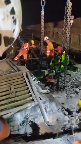 Four individuals, appearing to be construction workers, are visible in the video. They are dressed in safety gear, including hard hats and high-visibility vests. Two of the individuals are in orange vests and white hard hats, one in a green vest and pink hard hat, and another in a yellow hard hat and orange vest. The scene appears to be a construction site at night, with artificial lighting present. There are large metal chains hanging from overhead structures, suggesting the use of heavy machinery for lifting or demolition. Debris, including concrete and metal, is scattered around the ground. One person is using a flashlight to illuminate an area on the ground. The workers seem to be engaged in some form of repair or demolition work.