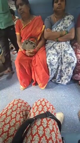 Two women are sitting on a blue bench, possibly on a train. The woman on the left is wearing an orange sari and has her eyes closed. She is holding a purse on her lap. The woman on the right is wearing a blue and white sari and has her arms crossed. Her eyes are open. In the foreground, a person's legs are visible, wearing red and white patterned pants and a black backpack strap across their lap. The floor is blue and speckled. Other passengers are visible in the background.