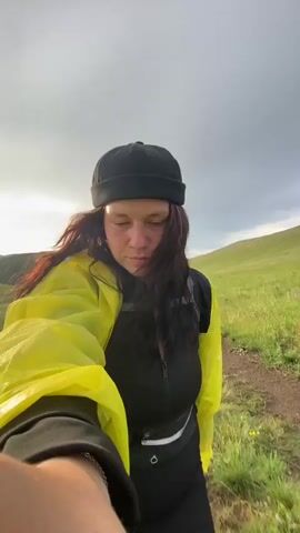 A person wearing a yellow raincoat and a black beanie is standing in a grassy field, holding a camera out to record themselves. The person is smiling at the camera, and their hair is long and dark. The background consists of rolling green hills under a cloudy sky. The person appears to be hiking or exploring.