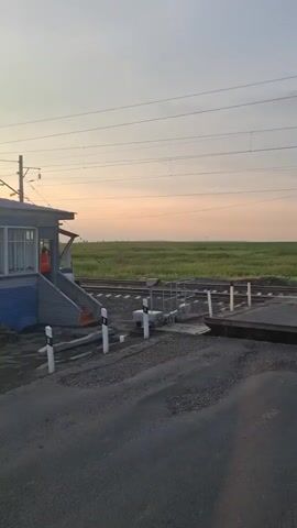 A train crosses a railroad track at a crossing. The train is gray and red with the letters "РЖД" on its side. The sky is overcast with a pale orange hue, suggesting either sunrise or sunset. In the foreground, there is an asphalt road with a railroad crossing gate. To the left, there is a small building with windows and a person in an orange vest standing on the steps. The crossing gate has red and white striped barriers. The train is moving from left to right across the frame.
