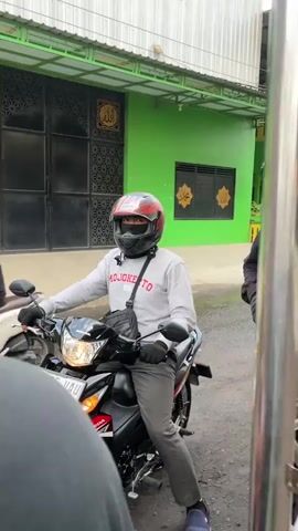 A person wearing a red helmet and a grey long-sleeved shirt with text "MOJOKERTO" is sitting on a black motorcycle, looking forward. The person is wearing black gloves and dark pants. A black crossbody bag is slung over their shoulder. The motorcycle is parked on a paved surface, possibly a street or alley. In the background, there is a bright green building with black window frames. Above the windows, there are decorative patterns and Arabic calligraphy. To the left, a large black metal gate is visible. The lighting suggests it is daytime. The scene appears to be outdoors in an urban or semi-urban environment. The angle of the shot is from the perspective of someone on another vehicle or in front of the motorcycle.