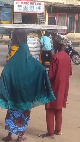 The video shows a street scene. In the foreground, a woman wearing a green hijab and a patterned skirt stands beside a man in a maroon outfit and a patterned hat. They are looking at a yellow auto-rickshaw (tuk-tuk) which is loaded with sacks.  Behind them is a white car and a black motorcycle parked on the side of the road.  Behind the tuk-tuk, a man in a light blue shirt is adjusting the sacks. A building with a sign "I.C. MARC INTL CO." is visible in the background. The street seems busy with cars and motorcycles passing by.
