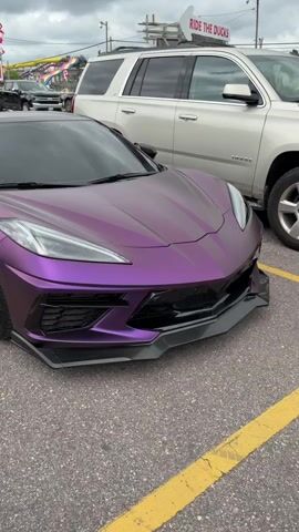 A close-up shot pans across the front driver's side wheel and fender of a matte purple Chevrolet Corvette C8. The car is parked in an asphalt lot with yellow painted lines visible. In the background, a white Chevrolet Suburban is parked, and further behind it, a sign with "RIDE THE DUCKS" written on it is visible. The car's tires are black with bronze multi-spoke rims.