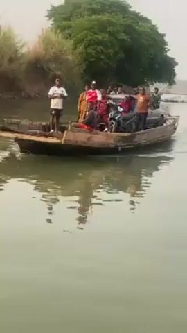 A wide shot captures a wooden boat loaded with people, a motorcycle, and a car navigating a river. Several individuals are standing on the boat, while one person is seated. The boat is positioned towards the center of the frame, moving slowly across the calm water. Lush greenery and trees line the riverbank on both sides. The overall lighting suggests it is daytime.