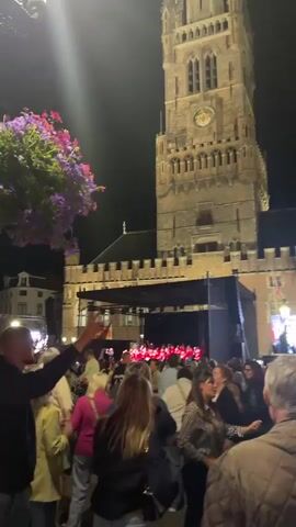 A nighttime outdoor gathering is taking place in front of a tall, old stone tower, possibly a belfry, adorned with a clock face. A stage is set up with a black backdrop, and a group of people in red outfits are visible on it, performing or singing, illuminated by purple and white stage lights. A large screen displaying an image is also set up on the right side of the stage. A crowd of people, diverse in age and gender, are gathered in the plaza, facing the stage and screen. Many are holding up their phones, likely to record the event. In the foreground, several individuals are captured from behind, with one person in a brown quilted jacket raising their hand. To the left, a large hanging planter filled with vibrant purple flowers adds a splash of color to the scene. The atmosphere appears festive, with people enjoying the performance. The ground is paved, and the surrounding buildings have architectural details consistent with historical European structures.