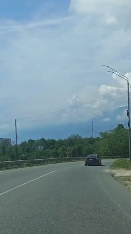 The video shows a street with cars driving on it, surrounded by lush green trees and mountains in the background. The sky is partially cloudy. There are utility poles with wires running along the side of the road. A green metal fence is visible behind the utility poles, separating the road from an area with more trees and possibly houses. The camera pans slowly to the right, revealing more of the mountainous landscape.