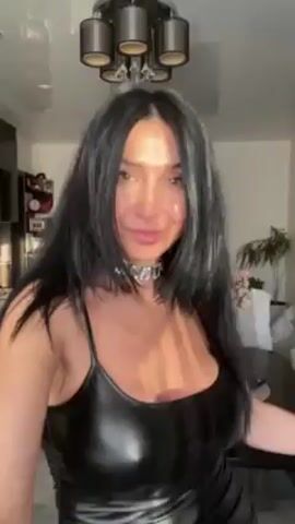 A woman with dark hair and light skin is standing indoors, smiling at the camera. She is wearing a shiny black dress with thin straps and a silver choker necklace. The room behind her includes a modern light fixture, a dark cabinet with decorative items, a dining table, and a glimpse of a plant in a vase. She appears to be making a short video or taking a photo in what seems to be her home.