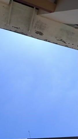 The video shows a clear blue sky from a low angle. A portion of a building is visible at the bottom of the frame, with windows and a dark roof. The top of the frame shows a concrete overhang with visible signs of wear and tear. The camera appears to be panning slightly upwards.
