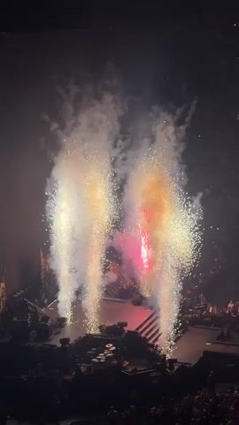 The video displays a concert or live event. The first few seconds show pyrotechnics, with bright white and pink sparks erupting upwards, obscuring the stage below with smoke and light. The scene then transitions to a wide shot of a darkened arena, illuminated by intense blue stage lighting. Silhouettes of audience members are visible in the foreground, their faces obscured, suggesting they are looking towards the stage which is not clearly visible. The overall atmosphere appears to be one of excitement and spectacle.