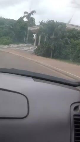 The video, shot from inside a vehicle, pans across a landscape featuring lush greenery and a contemporary architectural structure with a flat roof supported by columns. A paved pathway and steps lead up to the structure, which appears to be an entrance or pavilion. The sky is overcast. The camera movement is smooth, suggesting the vehicle is in motion.