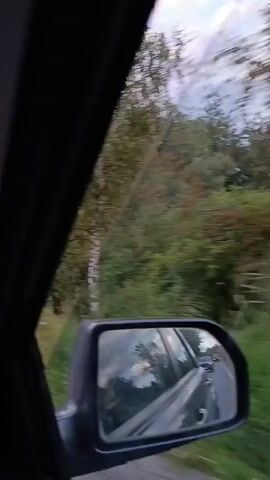 This is a first-person perspective video from a car driving down a rural road. The road is paved with a white line on the right side and a yellow line in the center. On the right side of the road, there is a dense line of trees and vegetation. On the left side of the road, there are more trees and some houses scattered throughout. The sky is cloudy with patches of sunlight breaking through, suggesting it is either early morning or late afternoon. There are a few other vehicles visible on the road ahead, including cars and a truck. The overall environment is rural and appears to be a typical countryside setting.