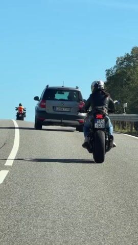 A motorcycle rider wearing a white helmet and a black jacket with blue jeans rides on an asphalt road under a clear blue sky. The rider is followed by a gray SUV and another motorcycle. Trees and foliage line the right side of the road, and a yellow road sign is visible in the distance. The motorcycle has a license plate that reads "CZ 965".