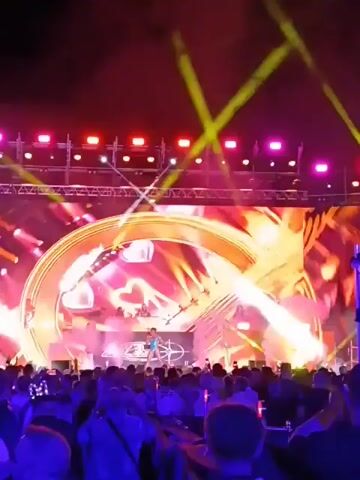 A large crowd is gathered in an outdoor space in front of a large stage. The stage is lit with colorful lights, including pink, white, yellow, and red. There is a large screen on the stage displaying various visuals, including a large circular shape and other geometric designs. A person is visible on the stage, presumably performing. Many people in the crowd are holding up their phones, likely recording the event. The scene is energetic and lively, with a party-like atmosphere.
